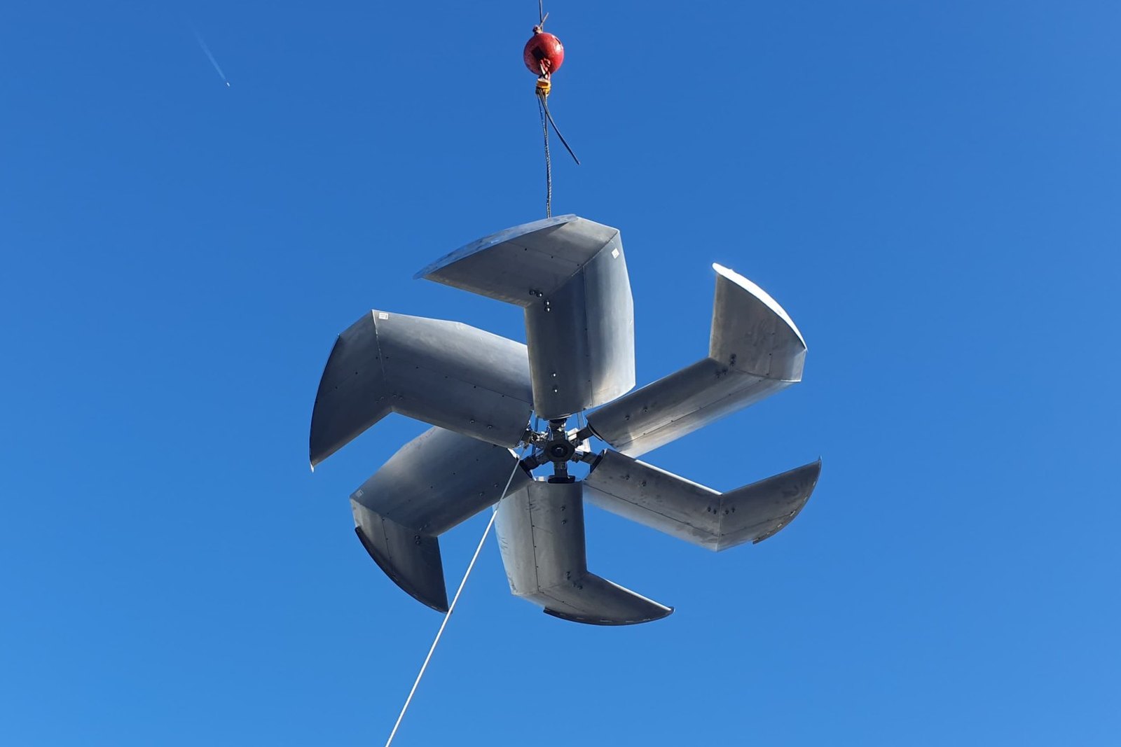 A five blade axial fan assembly hanging from a crane hook against a clear blue sky, the airfoil section of each blade visible
