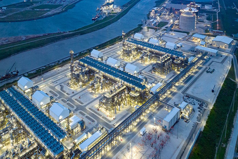 Aerial view at dusk of a large gas plant beside a ship channel, three long banks of air cooled heat exchanger fans lit along the process trains